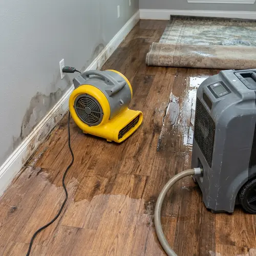 Air mover and dehumidifier drying a wet wood floor beside a stained wall, water and fire damage restoration